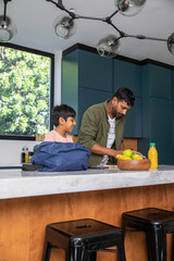 Indian father with son standing at marble kitchen island slicing fruit with knife beside backpack © wavebreak3