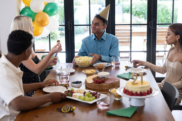 Diverse friends celebrating 21st birthday around dining table at home with cake, popcorn
