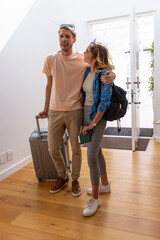 Couple standing in bright entryway holding rolling suitcase and passport while wearing backpack