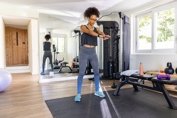 African American woman in her 30s holding cable machine handles standing on mat at home gym
