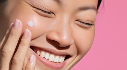A smiling Asian woman with closed eyes applies a dab of white cream to her cheek.