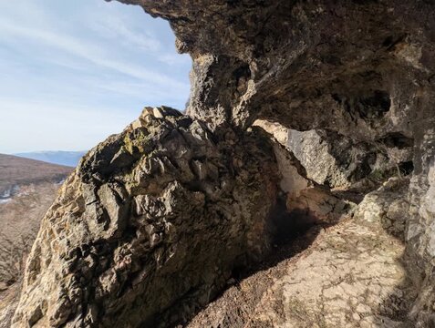 Vista tra le pareti frastagliate, rivelando giochi di luce e ombra che esaltano la texture della pietra. Sullo sfondo, un panorama naturale si apre con colline e foreste che si perdono all&rsquo;orizzonte