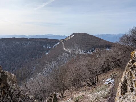 La scena si apre su un crinale montano immerso nella quiete invernale. Il sentiero corre lungo la dorsale, alternando tratti rocciosi a zone dove la neve resiste ancora al sole