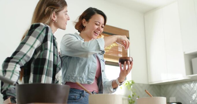 Diverse female friends at home counter, woman sprinkling salt from wood bowl into bowls for dinner