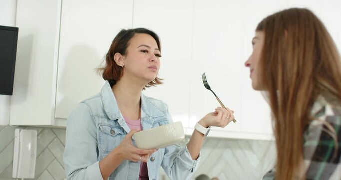 Two women chatting in kitchen, woman tasting beige bowl while holding fork and friend showing phone