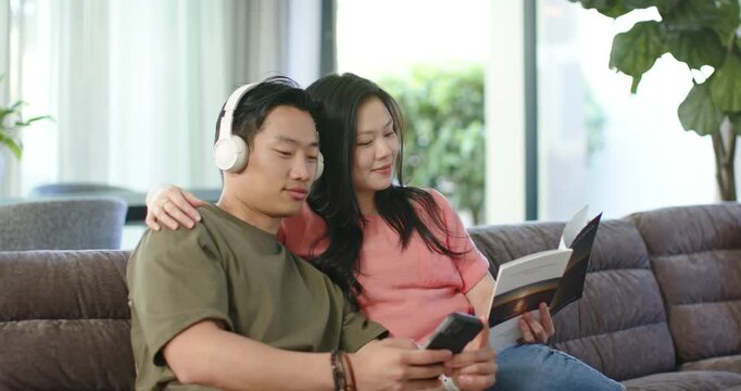 Diverse couple on couch, man wearing headphones scrolling phone, woman leaning, comparing booklet