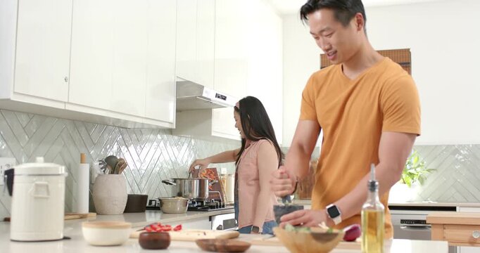 Diverse couple making dinner at home, woman tilting oil bottle into pot, urging man grinding spices