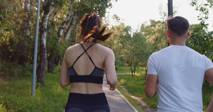 Fit couple of athletes running marathon together in morning in summer park. Back view of sporty woman and man runners in sportswear jogging on walking track to enjoy fresh air and endurance training