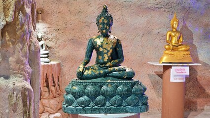 Obraz premium CHONBURI, THAILAND – NOVEMBER 29, 2025: A dark green statue of the Buddha in a meditative pose is covered in gold leaf within an artificial grotto at Wat Bot temple.