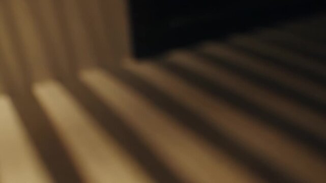 Sunlight casting strong, angled parallel lines of light and shadow onto a light colored surface, creating a minimalist graphic pattern with warm tones and soft focus