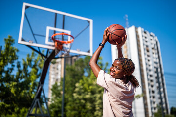 Young African American woman taking a basketball shot on an urban outdoor court during a sunny summer day.