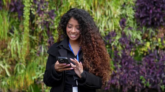 Young smiling woman with long curly hair using her mobile phone. Businesswoman texting and scrolling on her smartphone outdoors