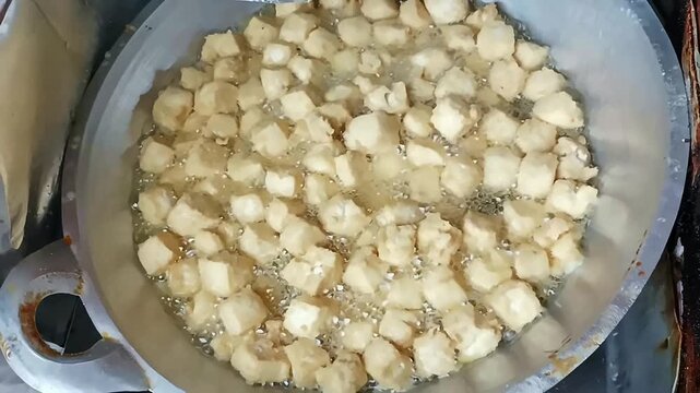 Deep-frying small tofu cubes in hot bubbling oil at a street food stall. Traditional cooking process of Indonesian crispy Tahu Goreng snacks in a large metal wok.
