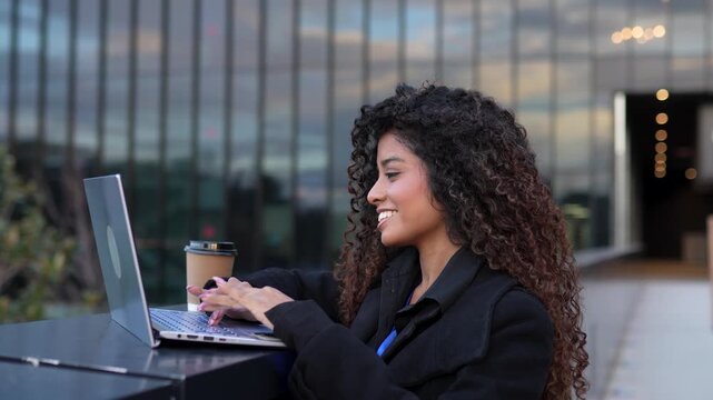 Beautiful young professional woman with curly hair smiling and typing on her laptop computer, working remotely in a modern urban city plaza