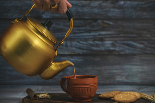 A person is pouring masala karak tea/chai into a cup from a gold teapot. The scene is set on a wooden table with a plate of cookies. Scene is calm and relaxing, as the person pours the tea