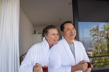 Diverse senior couple wearing bathrobes standing on balcony overlooking ocean sipping tea