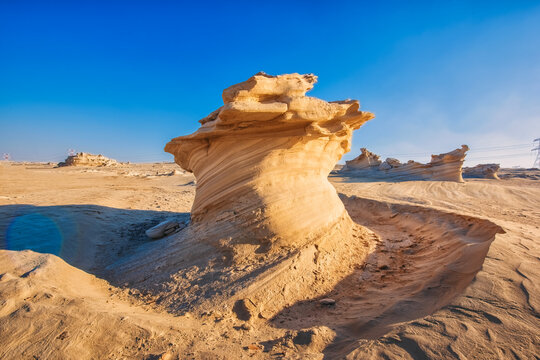 A large rock formation sits in the desert, surrounded by a vast expanse of sand. The sky is clear and blue, and the sun is shining brightly. The scene is peaceful and serene