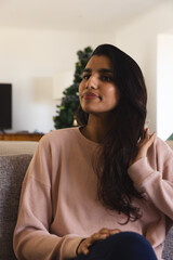 Asian woman sitting on gray sofa in living room wearing pink knit sweater by Christmas tree