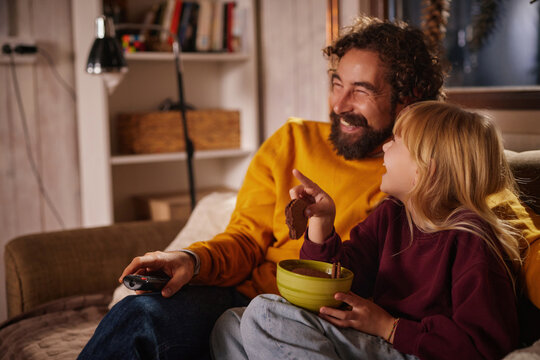 A man and a young girl are seated comfortably on a couch, watching a movie together and sharing joyful moments while eating popcorn and cookies.