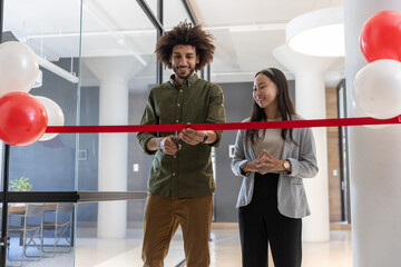 Diverse coworkers cutting red ribbon at office lobby with glass partitions, balloons, scissors © wavebreak3