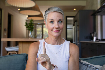 Senior woman sitting on sofa in kitchen by island with pendant lamps, raising index finger © wavebreak3