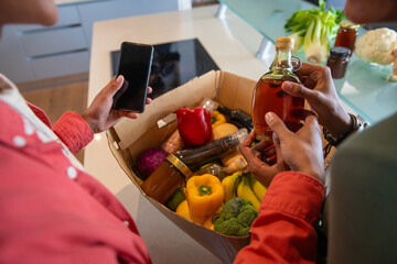 Diverse couple holding smartphone over produce box checking oil bottle at kitchen island © wavebreak3
