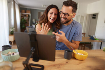A happy couple joyfully shows off an engagement ring during a video call while enjoying breakfast...