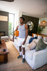 Naklejka premium African American man holding basketball while posing in modern living room with wooden coffee table