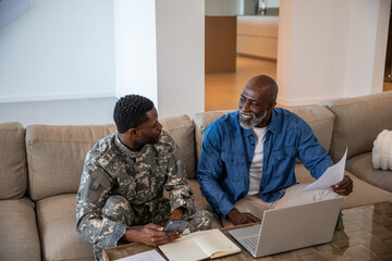 Naklejka premium African american father, son in camouflage uniform reviewing documents on laptop in living room