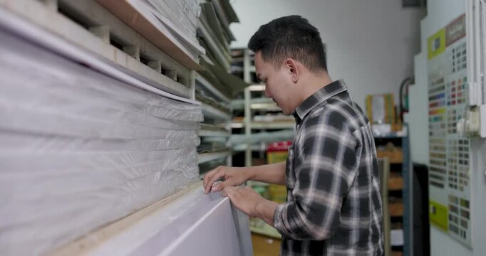 Asian adult man carpenter walks along rows, sliding hands across stacked wood boards, inspecting surface condition, recording inventory levels, managing wood material selection for furniture making