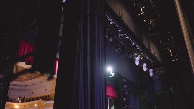 Dark backstage view of theatrical scenery and lighting equipment descending. On the left, the theater hall with balconies and red curtains is visible