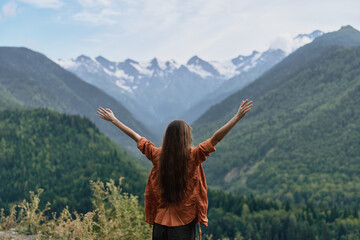 Obraz premium Woman with arms raised in a vast mountain landscape, embracing nature and freedom, gazing over green valleys toward snowy peaks under a clear alpine sky