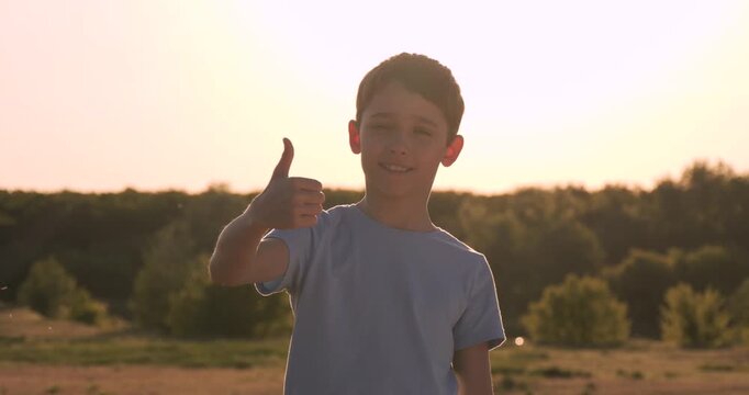 Child boy thumbs up smiles at sunset in summer park. Kid boy thumbs up stands confident. Warm golden light paints sky. Outdoor success. Child boy thumbs up shows approval motivation, positive mindset.