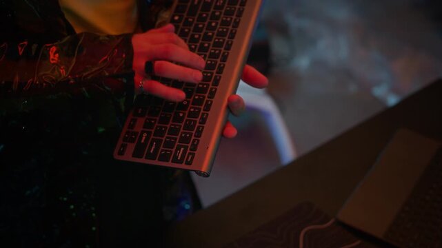 Tilt up shot of Caucasian man in virtual reality visor and black reflective jacket using keyboard while decrypting data on computer system in cyberpunk neon room