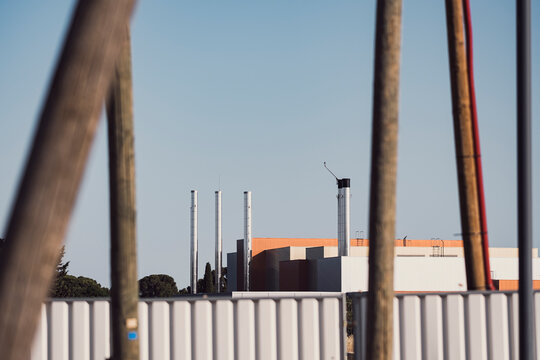 Factory industrial chimney construction fence perspective on structure framework under sky with facility seen through foreground posts creating depth