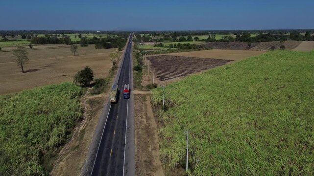 Aerial highway road through rural farmland agriculture field and crop landscape, drone view of long straight asphalt route with truck traffic, green patches and dry plowed land under clear summer sky