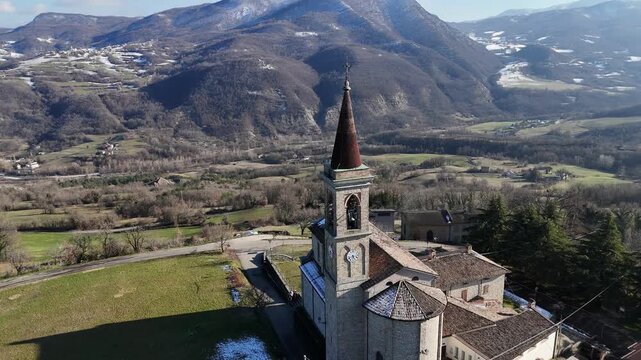 Drone aerial of Chiesa dei Santi Salvatore e Gallo in I Rabbini, Morfasso, Piacenza, Italy, showing a stone rural church with spire and clock tower above the rolling Val d&rsquo;Arda hills of Apennines