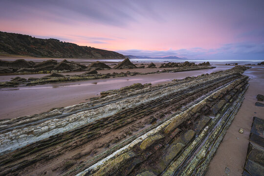 View of layered rock formations stretch across the sandy beach under a pastel sky, creating a surreal landscape of nature's artistry, Ugeraga, Basque Country, Spain.