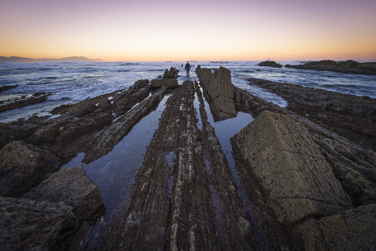 View of layered rock formations stretch into the sea under a vibrant sunset sky, with a lone figure standing in the distance, Ugeraga, Basque Country, Spain.