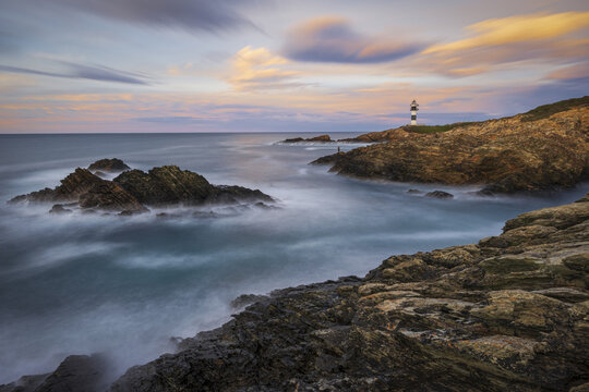 View of the lighthouse standing tall on the rocky coast, the ocean waves crashing gently, and the sky painted with soft hues of dawn, Ribadeo, Lugo, Spain.