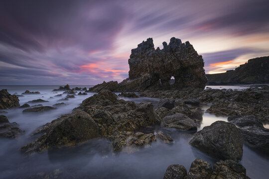 View of jagged rocks rise defiantly from the swirling, milky sea under a vibrant sky ablaze with hues of pink, purple, and orange, Luarca, Spain.