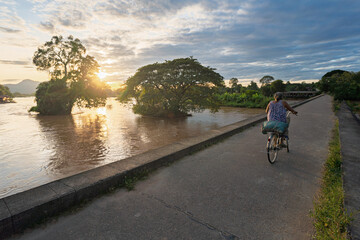Naklejka premium A person cycling along a riverside path at sunset in Don Det, Laos, with calm water, trees, and warm evening light.
