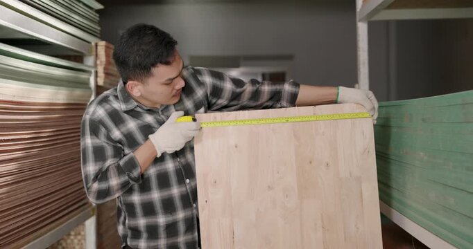 Asian male woodworking business owner stretches tape measure across wooden panel inside material store, verifying width dimension and material consistency for interior furniture production workflow