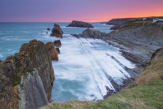 View of the rugged coastline where the turquoise sea crashes against the stratified rocks under a vibrant twilight sky, Santander, Cantabria, Spain.