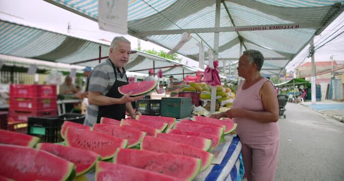 Elderly market vendor and customer talking across watermelon display at outdoor stall, everyday shopping moment, human connection, local market routine