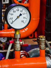 Pressure gauge on a bright orange pipe in an industrial setting with various equipment in the background during daytime