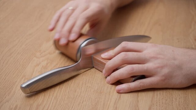 Hands sharpening steel kitchen chef knife using rolling sharpener on wooden table. Maintenance of blade edge for culinary cooking with metal abrasive sound of sharpening tool.