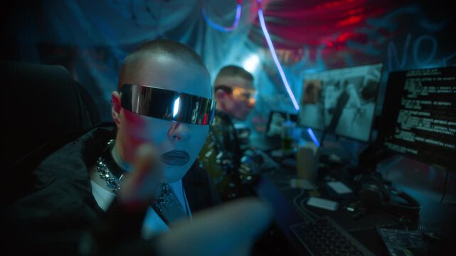 Medium close up shot of woman with shaved head wearing futuristic reflective glasses typing on invisible virtual keyboard in front of camera, working with man analyzing footage and coding on computers