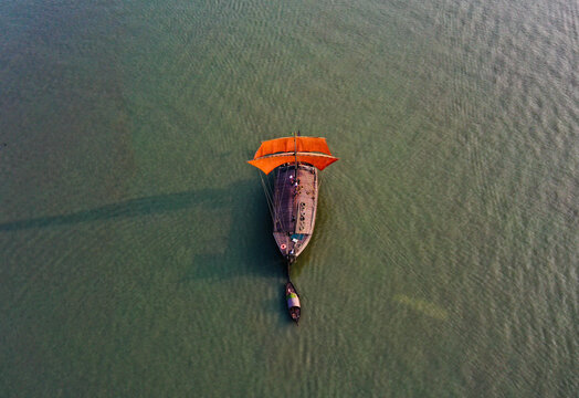 Pabna, Bangladesh - 16 December 2020: Aerial view of a traditional wooden boat with vibrant orange sails cutting through the serene, rippled waters of the Padma River.