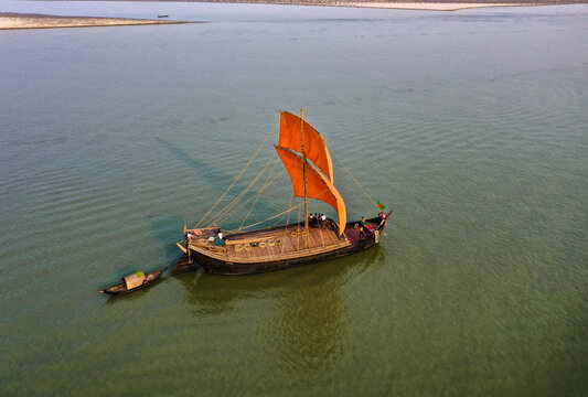 Pabna, Bangladesh - 16 December 2020: Aerial view of a traditional wooden boat with a vibrant orange sail glides along the calm, reflective waters of Padma river.